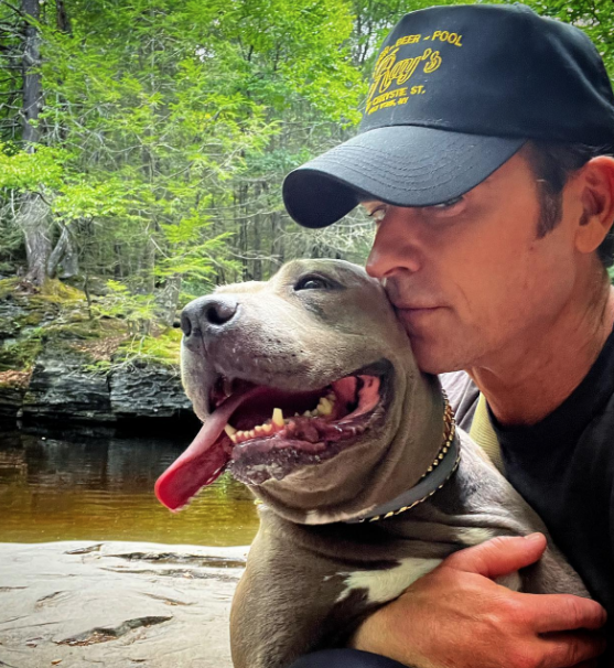 Justin Theroux hugs his grey pit bull with a river in the back