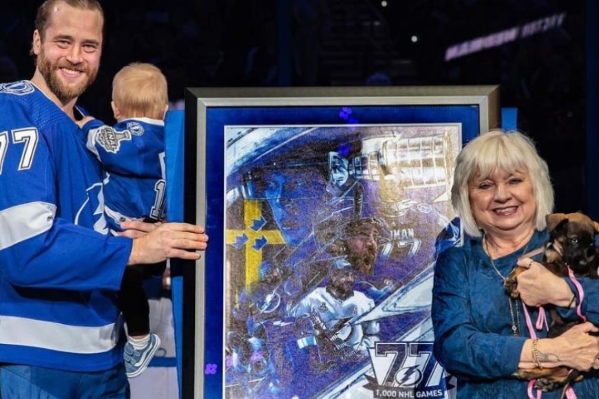 A Tampa Bay Lightning player Victor Hedman, is smiling at the camera holding his son, and Sherry Silk, HSTB CEO is holding a dark brown puppy and smiling into the camera. In between the people sits a large framed collage of Victor Hedman.